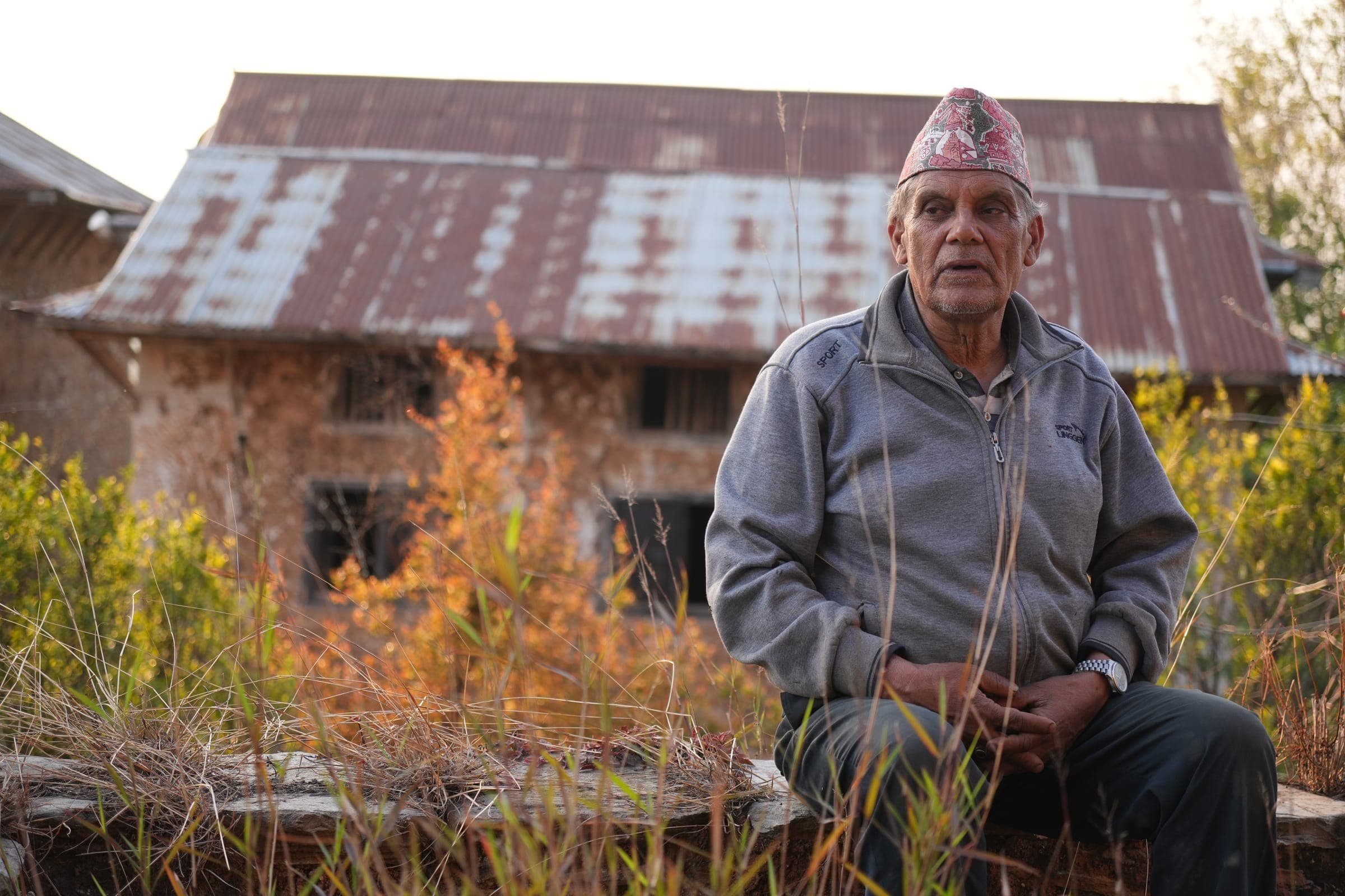 An elder of the village in a dhaka topi, sitting in front of an old stone house